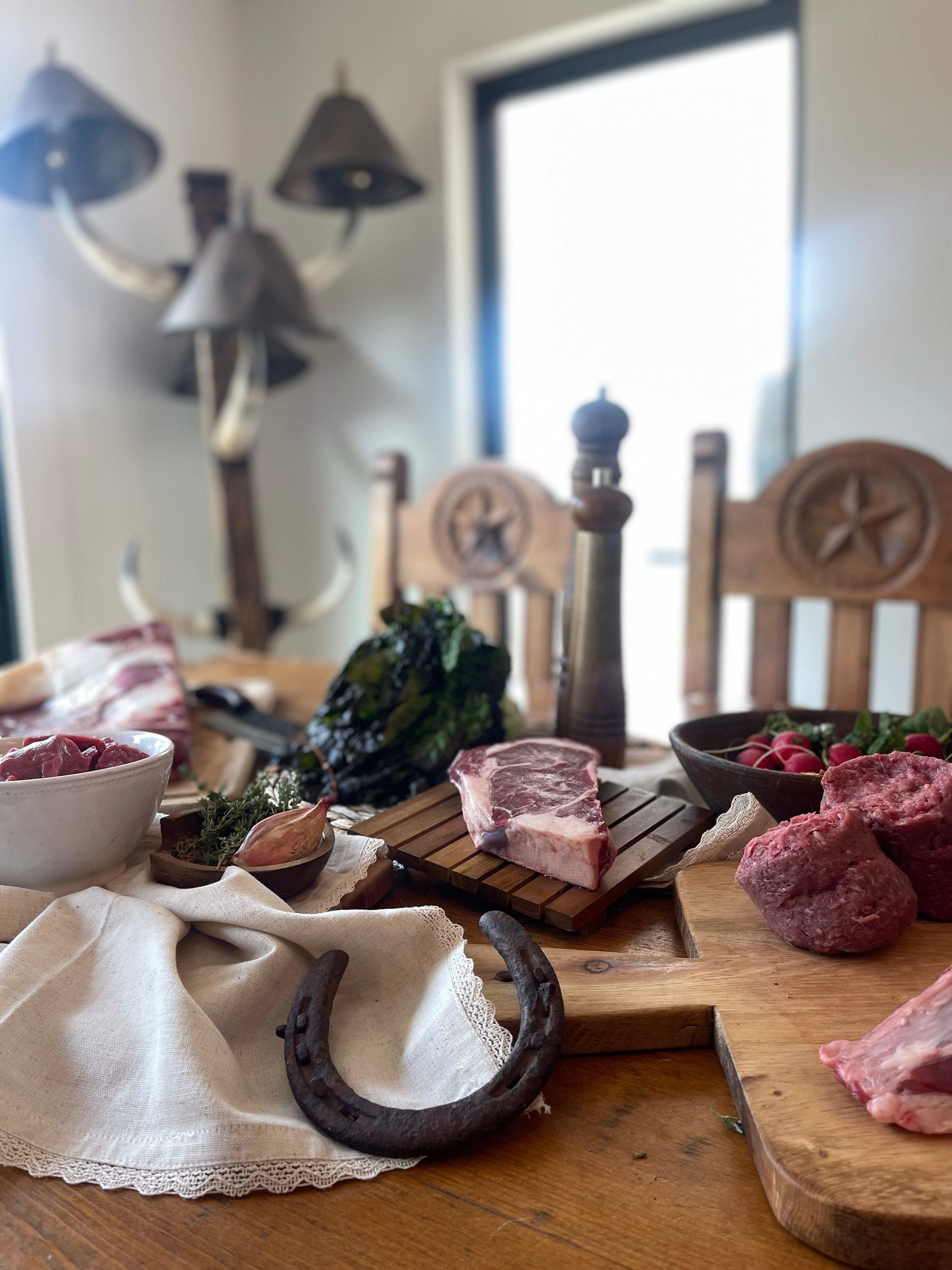Raw meat cuts, bowls, and rustic kitchen items arranged on a wooden table in a home setting, with chairs and a window in the background.